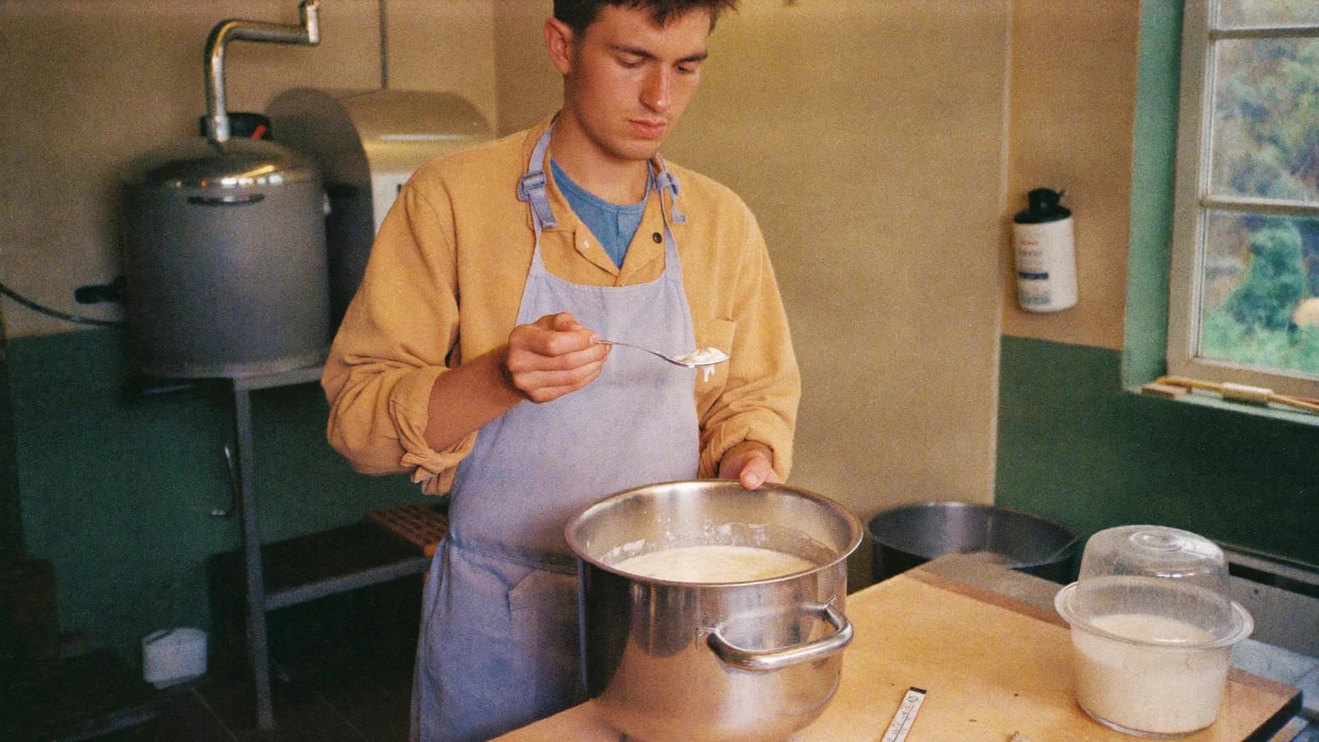 Dairy artisan checking cultured milk beside a stainless-steel pot.
